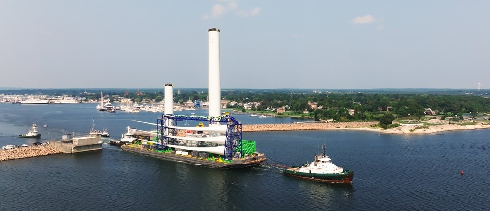 Marine vessel carrying wind turbine tower components and blades heading out to sea with aid of tug boats