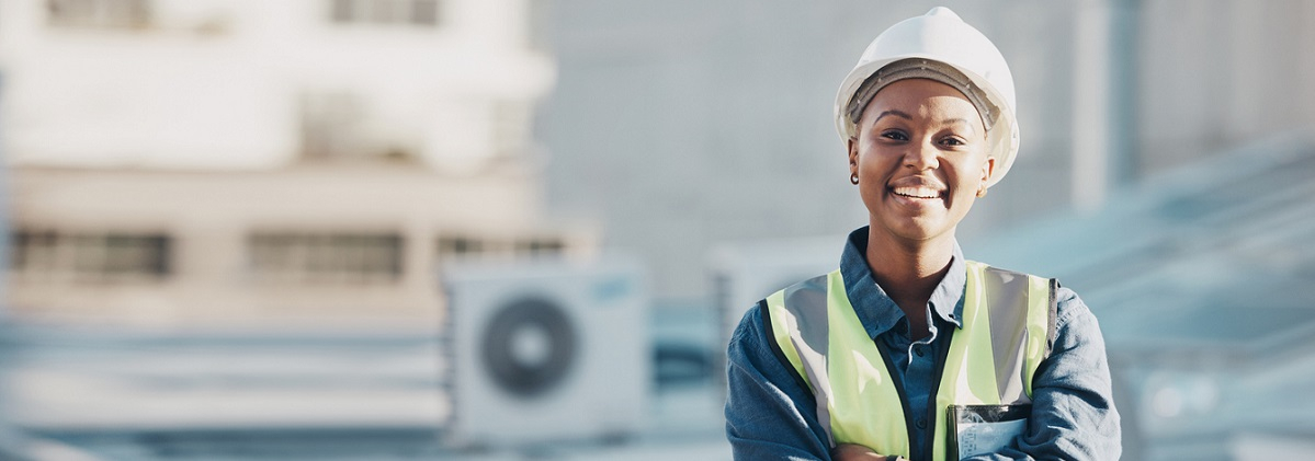 Smiling young black woman in hard hat and safety vest on roof with heat pump in background